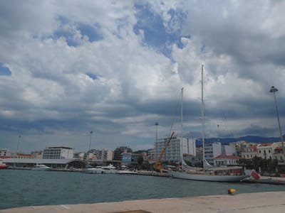 The image depicts a waterfront scene in the Municipal Unit of Patras, Greece. In the foreground, a concrete pier runs along the bottom of the frame. The water is a deep blue-green and appears choppy. Moored along the pier are several yachts of various sizes. A large sailboat with tall masts dominates the right side of the frame, with a red and white flag attached to its stern. Behind the yachts, a cityscape unfolds. Buildings of different architectural styles and heights line the waterfront, with some appearing to be modern apartment buildings and others older structures with red-tiled roofs. Several tall light poles are visible throughout the scene. The sky is filled with dramatic, dark, and light gray clouds, with patches of blue sky visible in between. A yellow construction crane is positioned behind some of the buildings, suggesting ongoing construction or renovation work. The overall impression is one of a busy port or marina on a cloudy day.