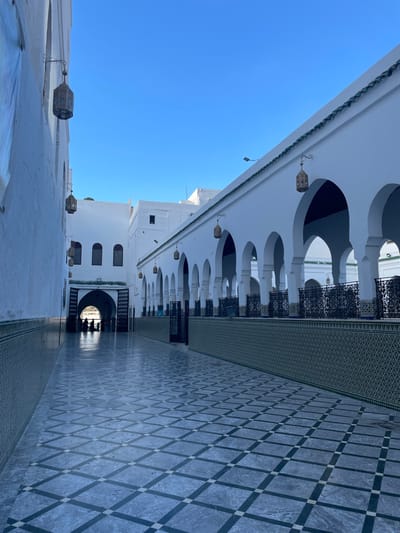 This image captures a long, outdoor paved corridor within a building complex, identified as being in Moulay Idriss Zerhoun, Morocco. The corridor is bordered by white-painted structures under a clear blue sky.
On the left, a solid white wall extends into the distance, featuring a lower section adorned with green and white geometric tiles. Several ornate, brass-colored lanterns are suspended from dark, scrolled brackets along this wall. A portion of a light blue banner or fabric is visible near the top left corner of this wall.
On the right, a parallel white structure displays a series of evenly spaced horseshoe arches, forming an open colonnade or walkway. A narrow, green-tiled roofline runs along the top of this arched structure. Below the arches, a decorative barrier is present, consisting of dark, wrought-iron grilles atop a wall section also patterned with green and white geometric tiles, matching the left side. Multiple brass-colored lanterns similar to those on the left wall hang within the arched walkway.
The floor of the corridor is paved with large, grey square tiles, arranged in a diagonal pattern with smaller white squares at their intersections, creating a leading line towards the background.
At the far end of the corridor, a large, dark arched entrance leads into an interior space. Approximately three to five indistinct human figures are visible in silhouette within or immediately beyond this archway, appearing to be standing or moving. Beyond the arch, a brightly lit area suggests another open space or courtyard. Additional white buildings with dark arched windows are visible in the distant background. The scene is illuminated by natural sunlight, casting subtle shadows.