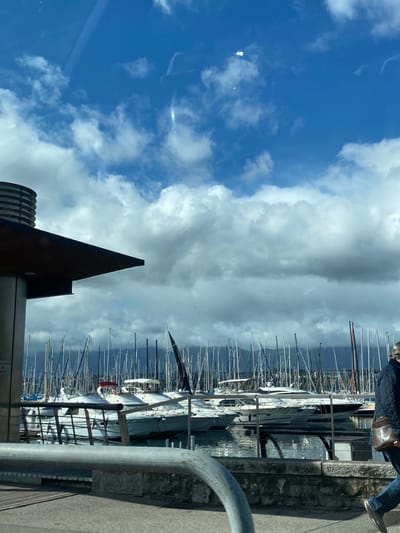 The image captures a marina scene in Cologny, Switzerland, under a sky exhibiting both clear blue patches and extensive white and grey cumulus cloud formations. A notable glare or reflection is visible in the upper central sky area.
The mid-ground is dominated by a marina filled with numerous docked boats, predominantly sailboats, characterized by a dense arrangement of their vertical masts. Several white motor yachts are also present among the sailboats. The water within the marina displays reflections of the vessels and sky. In the distant background, a dark, undulating mountain range is visible beneath the clouds.
In the foreground, on the left, a portion of a modern structure is visible, featuring a dark, flat overhang and a light-colored support pillar. A cylindrical, louvered ventilation unit is positioned on top of the structure's roof. A curved, light-grey metal handrail traverses the lower-mid section of the frame, positioned in front of a stone or concrete retaining wall that borders the marina. On the right side of the foreground, a person is depicted in mid-stride, walking from right to left. The individual is wearing dark blue jeans, a dark quilted jacket, and carries a brown leather satchel slung over their right shoulder. Their head and upper torso are outside the frame. They are walking on a paved surface adjacent to the marina.