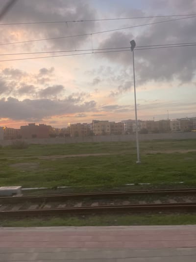 A sideways view captures a cityscape at dusk. Buildings line the horizon under a cloudy sky with hues of orange. Power lines stretch across the sky. A grassy field extends along the right side, adjacent to railway tracks. A platform edge is visible on the far right.