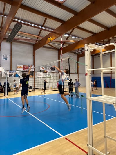 The image displays an indoor volleyball game or practice session occurring within a sports hall in Sennecey-le-Grand, France. In the foreground, two male players are captured airborne on opposite sides of a black volleyball net. The player on the left wears a dark blue jersey with "SNC" and the number "8" on the back; he is positioned with arms raised, appearing to prepare for a spike or block. The player on the right, in a light-colored patterned jersey, is also mid-air with his left arm extended over the net, in the act of contacting a yellow and blue volleyball which is visible high above the net.
The playing surface is a blue synthetic court marked with white boundary lines. A white and red striped net antenna is visible on the net's right side, next to a partially visible white metal referee stand. In the background, multiple other individuals are present, some appearing to be additional players, while others observe the activity from the sidelines. The sports hall features light-colored walls, a high ceiling composed of exposed laminated timber beams and corrugated metal panels, and overhead rectangular light fixtures and heaters. Advertising banners, including one labeled "VBS" and another for "Netto," are affixed to the distant walls, along with a wall-mounted basketball hoop.
