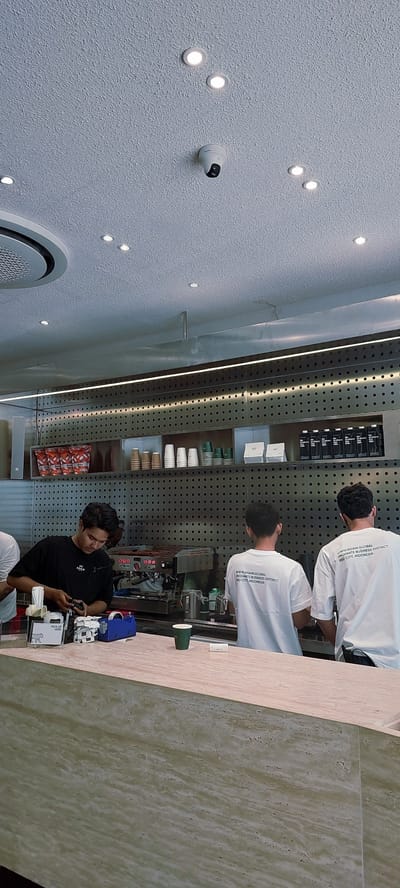 In the city of Lengkong Kulon, Indonesia, a coffee shop scene unfolds with a focus on the bar area. Three individuals are visible, two of whom appear to be baristas working behind a counter. One barista, wearing a black t-shirt, is actively engaged with an espresso machine, holding a portafilter. To his right, another individual in a white t-shirt is also behind the counter, facing away from the camera, suggesting they are also an employee. A third person, also in a white t-shirt with text on the back, stands further to the right, also facing away. The counter itself is made of a light-colored, textured material. Behind the baristas, shelves are stocked with various items including bags of coffee, stacks of cups, and boxes of milk cartons. The ceiling features recessed lighting and a security camera. The overall environment suggests a modern and functional coffee shop setting.