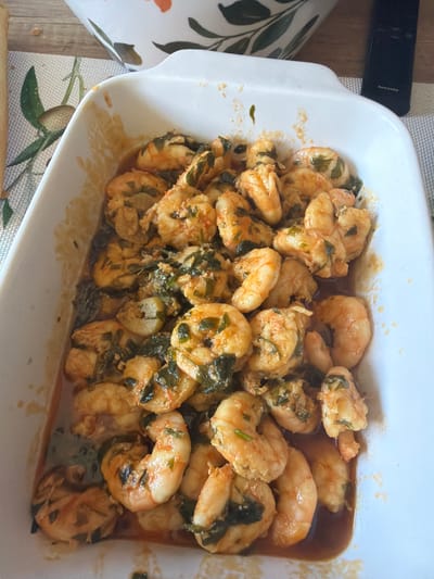 A top-down, close-up shot reveals a white, rectangular ceramic baking dish filled with cooked shrimp in a sauce, likely on a patterned tablecloth or placemat. The shrimp are predominantly pink and orange, mixed with chopped green herbs (possibly parsley or cilantro). A dark sauce coats the shrimp, with pooling visible in the bottom of the dish. In the upper left corner, a black Samsung remote control partially enters the frame. Based on the image's origin, the scene is likely located in Azuqueca de Henares, Spain. No people are visible. The focus is on the prepared dish of shrimp.