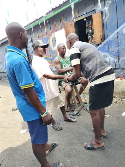Four adult males are depicted on a paved surface in Oworonshoki, Nigeria, against a building with corrugated metal siding. The leftmost man, partially visible and facing right, wears a blue polo shirt with yellow trim, dark blue shorts, and black slides, with a light blue wristband on his right wrist. To his right, a man wearing a dark baseball cap, white t-shirt, and light shorts is shaking hands with a seated man. The seated man, wearing a green Boston Celtics jersey (number 34), dark shorts, and dark slides, sits on a wooden stool and wears a gold-colored chain. A fourth man, seen from the back, wears a grey vest over a horizontal striped shirt, black shorts, and black slides, and is also shaking the seated man's hand. The background building features multi-colored graffiti, an open window or doorway revealing patterned fabric, and a large blue fishing net draped on the right side. Green and white triangular pennants are strung above the scene. The ground is a worn asphalt or concrete surface, and a red bucket is visible near the fishing net. All individuals are wearing open-toed slides or sandals.