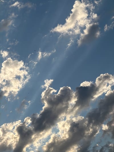 A vertical, eye-level, medium shot captures a dramatic sky filled with clouds. The clouds are varied in color, ranging from bright white, illuminated by sunlight, to dark gray, suggesting depth and shadow. Wispy, lighter clouds are scattered throughout, adding texture to the scene. Rays of light pierce through the gaps in the clouds, creating visible beams against the clear blue sky. In the upper portion of the image, two small, dark objects, possibly birds or insects, are visible against the blue expanse. The lower portion of the image is dominated by heavier, more defined cloud formations. The image does not contain any identifiable landmarks or features to indicate a specific location within Arcahueja, Spain.