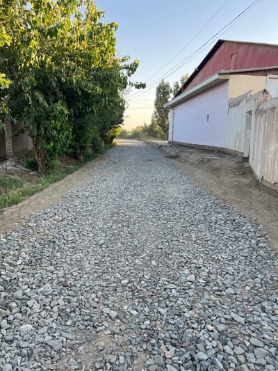The image depicts a street under construction in Hazorasp, Uzbekistan. The road is covered with a layer of gravel, indicating that it is not yet paved. On the left side of the street, there is a lush green tree with dense foliage. Adjacent to the tree, a portion of a building constructed with bricks is visible. On the right side of the street, a light purple building with a red roof stands, with its wall extending along the side of the road. A concrete gutter runs along the edge of the building. The sky is clear and blue, suggesting it is daytime. The scene appears quiet and devoid of any people or ongoing activity.