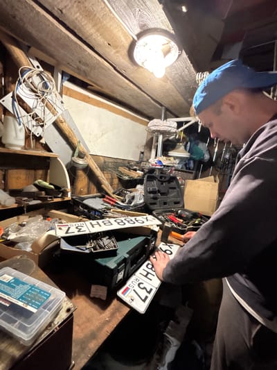 A man wearing a blue baseball cap and dark long-sleeved shirt works in a cluttered workshop. He is standing at a workbench, holding what appear to be Russian license plates. The workshop is dimly lit by an overhead lamp and filled with various tools, boxes, and miscellaneous items. The walls and ceiling are made of wood, suggesting a rustic or older structure. The overall impression is one of a working space dedicated to manual tasks, likely in a rural or semi-rural setting such as Dunilovo, Russia.