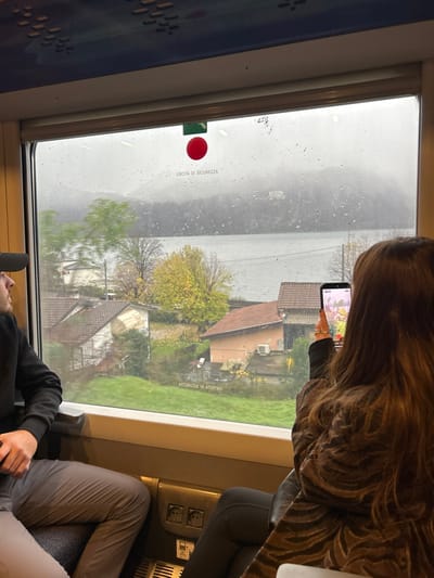 The scene depicts the interior of a train carriage looking out a window onto a landscape identified as Mandello del Lario, Italy. The weather is overcast with visible raindrops on the windowpane, suggesting recent or ongoing rain. The season appears to be autumn, characterized by trees with bright yellow foliage among greener ones.
In the foreground, inside the train, two individuals are visible. On the left, a male in a dark cap and dark long-sleeved shirt is seated, facing left and looking out the window. His hands are clasped. On the right, a female is seen from behind, with long brown hair, wearing a patterned brown and black jacket. She holds a black smartphone vertically with both hands, aiming it outwards, with the screen displaying a brightly colored image, indicating she is actively photographing or videoing the exterior scene. Multiple rings are visible on her right hand. Below the window, a dark grey panel with two European power outlets and a USB port is present. A blue patterned panel lines the ceiling of the train.
Outside the window, a view of Lake Como dominates the middle ground. Beyond the lake, tree-covered hills or mountains rise, partially obscured by the overcast sky. A small, light-colored building is faintly visible on a distant hillside. In the immediate foreground outside the train, several houses with terracotta-tiled roofs and light-colored walls are nestled on a grassy slope, interspersed with trees, descending towards the lake. One house features an external air conditioning unit. A red circular emergency handle with a green base, labeled "USCITA DI SICUREZZA" (Emergency Exit), is affixed to the top center of the window frame.