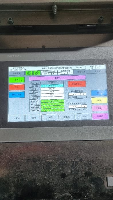 The image displays an active digital control panel screen, likely part of industrial machinery. The screen, housed within a gray metal frame, presents a graphical user interface primarily in Chinese characters with embedded numerical data. Key visible text identifies the system as "Xujing Inner Energy Chemical Industry SZN Sintering Machine Electronic Control System" and includes a date and time stamp of "2029-01-18 18:15:05." Various color-coded buttons are present, such as green for "Start/Activate" and red for "Stop," along with data fields related to "Winding Machine" and "Winding Speed Control." The surrounding hardware above and below the screen exhibits industrial wear, dirt, and metallic fasteners, indicative of an operational factory or control room environment. No individuals are visible. The textual content on the screen, particularly identifying Chinese entities, suggests the equipment is of Chinese origin or operated within a Chinese-speaking environment. The specific city of Kagan, Uzbekistan, is not discernible from the visual information provided in the image.
