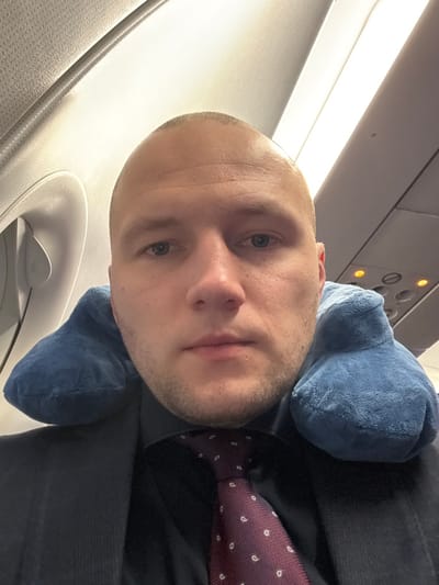A single adult male is depicted in a selfie-style portrait within the interior of a commercial aircraft cabin, located at Yemelyanovo Airport, Russia. The subject, appearing Caucasian, possesses a shaved head, blue eyes, and light facial stubble. He is dressed in a dark suit jacket, a dark collared shirt, and a burgundy tie featuring a white paisley pattern. A blue U-shaped travel neck pillow is worn around his neck. He maintains a neutral expression, looking directly forward. The background shows typical aircraft cabin architecture, including white overhead panels and a segment of an overhead console displaying illuminated orange indicator lights.