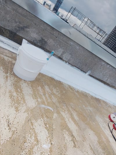 The scene depicts a concrete rooftop or elevated platform, situated in the city of Agios Tychonos, Cyprus, undergoing a waterproofing or painting application. A white plastic five-gallon bucket, containing a blue-handled brush, rests on the textured, light tan concrete surface in the lower left quadrant. A linear, approximately 15-centimeter wide band of freshly applied white waterproofing compound extends horizontally from left-center to right-center, along the base of a darker grey concrete wall. The concrete surface exhibits scattered white paint or compound splatter marks. The background reveals a distant urban skyline under a heavy grey sky, characterized by multiple high-rise structures, including buildings under construction with visible scaffolding and exposed concrete frameworks. A section of a metal barrier or scaffolding is present along the upper edge of the concrete wall. Two red-and-white spools on a black frame are partially visible in the bottom right corner. No human figures are present.