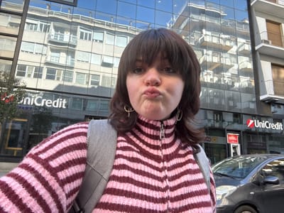 A young adult female stands outdoors, centered in a selfie-style photograph, with her lips pursed in a pouting expression, looking directly at the camera. She has dark brown, medium-length hair with bangs and a nose ring on her left nostril. She is wearing a pink and dark red horizontally striped knit sweater with a silver half-zip collar and silver earrings. A light gray backpack is visible over her right shoulder. The background consists of a modern building with a highly reflective glass facade, clearly displaying multiple "UniCredit" bank logos. This building is identified as the UniCredit branch located at Viale Michelangelo, 36, in Arezzo, Italy. The reflective glass facade shows a distorted reflection of an older building with balconies and windows, presumably across the street. To the lower right, a dark-colored hatchback car is partially visible, parked on the street. The scene is illuminated by natural daylight.