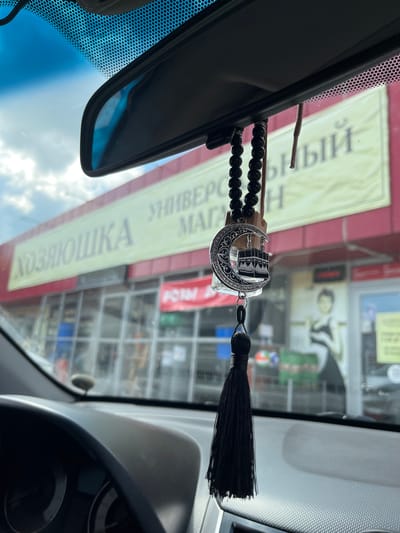 The image was taken from inside a vehicle, looking out through the windshield. In the foreground, a black tassel hangs from a string, attached to a keychain that features a crescent moon with an Islamic symbol of the Kaaba. Behind the keychain, out of focus, is a blurred yellow sign on a building that reads in Russian "ХОЗЯЮШКА" (Khozyayushka) and "УНИВЕРСАЛЬНЫЙ МАГАЗИН" (Universal Store). The building has large windows and a red awning. The sky is visible above the sign with some clouds. The dashboard of the car is visible in the lower part of the image. The location is specified as Novaya Otradovka, Russia.