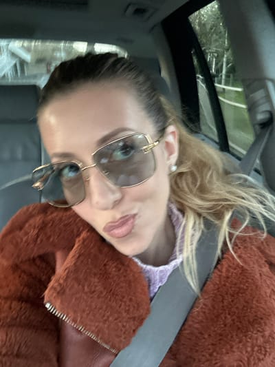 The image shows a person inside a car. The person is wearing sunglasses and a brown, textured jacket over a light purple top. They are wearing a seatbelt. The car appears to have a sunroof. The location is unknown, including the specific city of Basildon, United Kingdom.