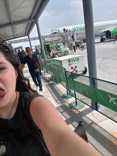 A rotated, eye-level outdoor shot shows a woman in the foreground, possibly taking a selfie. Her face, curly hair, black shirt and jewelry are partially visible. Behind her are other people, including a man in a black shirt looking towards the camera. In the background, a Viva Aerobus airplane sits on the tarmac, with passengers boarding via stairs. The boarding process and other airport personnel are visible. A Viva Aerobus branded luggage cart is also nearby. A green metal fence with airplane images runs along a walkway. The location appears to be an airport in Apodaca, Mexico, identified by the Viva Aerobus branding.