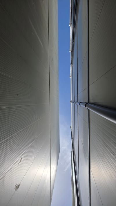 This media file captures a low-angle perspective looking upwards at the exterior of modern, large-scale buildings, likely warehouses or industrial facilities, under a clear blue sky. The buildings are constructed with corrugated, light-colored metal panels, creating a ribbed texture across their surfaces.

Two distinct building structures are visible, positioned in a way that they frame a central section of the sky. The upper structure appears closer to the camera and is seen from underneath, revealing its corrugated ceiling and a prominent grey pipe running horizontally across it, secured by black clamps. Below this, a thinner white pipe or conduit is also visible.

The lower structure dominates the bottom two-thirds of the image, its vast, ribbed surface extending upwards and seemingly towards the viewer. The bright sunlight reflects off these metal panels, creating shimmering highlights and emphasizing the geometric patterns. The sky above is a vibrant blue with a few wispy white clouds, suggesting a fair weather day.

There is no discernible activity or event taking place in the image. The scene is static, focusing on architectural elements and the expansive sky. The lighting indicates it is daytime, likely mid-morning or afternoon, given the brightness and the angle of the reflections. No people are visible. The location context provided is Moulins-sur-Orne, France, which hints at the type of industrial or agricultural infrastructure that might be found in such an area. No text is visible in the image.
