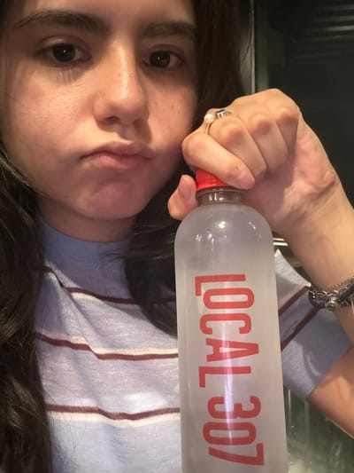 The image displays a female subject, cropped from the upper chest, holding a clear plastic bottle. Her dark hair frames her face, which features dark eyes and a neutral, slightly pursed expression. She wears a light blue, ribbed top with thin horizontal reddish-brown stripes. On her right ring finger, she has a silver-toned ring adorned with a white pearl and a darker inset stone. A multi-strand bracelet with various elements is visible on her right wrist. Her right hand grips the upper portion of the plastic bottle, which has a red screw cap. The bottle is partially frosted, indicating cold contents, and features the text "LOCAL 307" printed vertically in red. The liquid inside the bottle appears transparent. The background is uniformly dark and out of focus, providing no identifiable environmental details. The image contains no visual information to determine its location within Santiago, Mexico, or any other specific city.