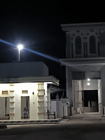 The photograph depicts a building exterior in Keudealue Ie Puteh, Indonesia, at night. The structure has a distinct architectural style, possibly religious or public, with white columns and archways. A streetlight shines brightly above the building, casting light on the facade. Two chairs are present near the entrance of a section of the building. A sign with directional arrows is visible near the building. The overall scene suggests a quiet, illuminated area within the city.