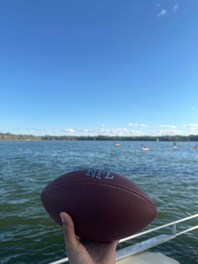 The scene depicts a recreational water environment near Micanopy, United States, viewed from a low vantage point, likely a boat or dock. In the immediate foreground, a light-skinned right hand firmly grasps a brown, pebbled American football. The silver letters "NFL" are clearly embossed on the side of the football facing the camera. A metallic railing, possibly part of a vessel or pier, is partially visible in the lower right corner, beneath the hand. Beyond the foreground, a large body of water, characterized by visible ripples, extends into the midground. Numerous individuals on various small watercraft, including kayaks or paddleboards, are scattered across the water in the distance, indicating recreational activity. The far horizon features a dense treeline, suggesting a natural shoreline. The sky above is largely clear and bright blue, with a few faint white clouds visible near the horizon. The overall lighting suggests a sunny daytime.