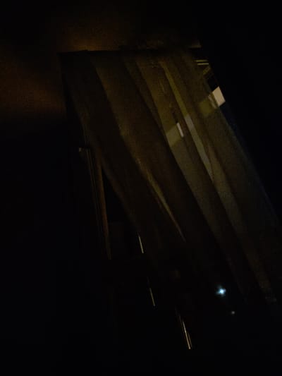 The image shows a dimly lit interior scene, possibly a room with a window covered by translucent curtains. The curtains are a pale yellow or cream color and hang in vertical folds, allowing some light to filter through. The light suggests it is nighttime outside, as there are some bright points of light visible, one of which is a noticeable blue-white glow. The overall impression is one of darkness and mystery. The specific location in Montrouge, France, is not discernible from the visual information provided in the image.
