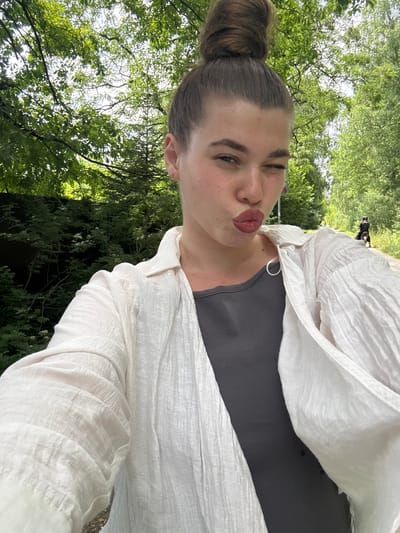 A young woman with her hair in a top knot is taking a selfie in an outdoor setting. She is wearing a white open shirt over a dark grey top and is puckering her lips. Behind her, lush green trees line a path, and in the distance, a person on a bicycle is visible. The scene suggests a pleasant day in a park or a trail, potentially in the city of Shevlyagino, Russia, given the context.