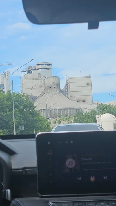 The image, taken from inside a vehicle, shows a large industrial complex, likely a cement plant, situated in Verkhnebakanskiy, Russia. The plant features numerous concrete silos and processing structures, some with visible pipework and ventilation shafts. A large, conical structure with a ribbed exterior dominates the center of the complex. Trees line the foreground, obscuring the base of some of the industrial buildings. A white truck trailer with a red stripe is visible to the right of the complex. The sky above is blue with scattered clouds. In the foreground, the dashboard of the vehicle is visible, including a dark touchscreen display showing a music playback interface with album art and song title. The overall scene depicts an industrial landscape on a clear day, observed from a moving vehicle.