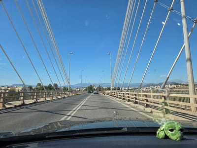 The image shows a view from a car driving across a modern cable-stayed bridge. The bridge has a light-colored road with white lane markings and a tan railing on either side. Several tall, white cables extend upwards from the road, disappearing into the bright blue sky. Streetlights are visible along the bridge. In the distance, there is a cityscape with buildings and green trees, nestled against rolling hills and mountains under a clear sky, suggesting a sunny day. The overall scene is one of transit and everyday movement. On the dashboard of the car in the foreground, a small, plush green turtle toy is visible. The Mercedes-Benz logo is also visible on the hood of the car. The time of day appears to be late morning or early afternoon, given the bright sunlight and the clear blue sky. The location context provided indicates the image was taken in Vasiliko, Greece.
