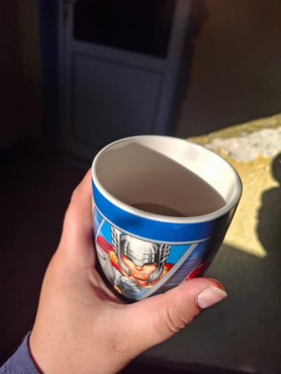 A hand holds a ceramic mug with an image of Thor on it. The mug is partially filled and sits on a textured, light-colored surface. The background is dark and out of focus, with a faint shadow cast on the right side of the image. The lighting suggests it is outdoors, possibly near a building.