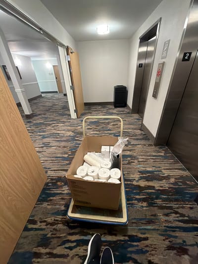 An overhead-oriented photograph captures a moment in a modern indoor hallway, likely within a hotel or commercial building. The scene is dominated by a blue and metal utility cart positioned on a patterned carpet, which features abstract streaks of blue, brown, grey, and off-white.

On the cart rests a large cardboard box filled with supplies. Visible inside are numerous rolls of white paper (resembling toilet paper or paper towels), stacks of white disposable cups, and a clear plastic bag containing more white items, possibly additional cups or containers.

In the background, two brushed metal elevator doors are visible. Above the top elevator door, a digital indicator displays the number "2," signifying the second floor. To the right of this, on the call button panel, there's a red rectangular button or sticker featuring the "Domino's" pizza logo. Another small gray plaque with an unidentifiable symbol and text is also present on the panel. A black, rectangular object, possibly a trash receptacle or speaker, sits on the floor near the elevators.

To the left, a light-colored wooden console table or counter stretches across the wall. Beyond this, a set of stairs with a metal railing and wooden treads descends. A wall-mounted light fixture illuminates the area near the stairs.

A person's feet, wearing dark shoes with white soles, are visible on the carpet next to the utility cart, suggesting they are in the process of pushing or pulling it. The overall impression is one of maintenance or restocking activity within a building.