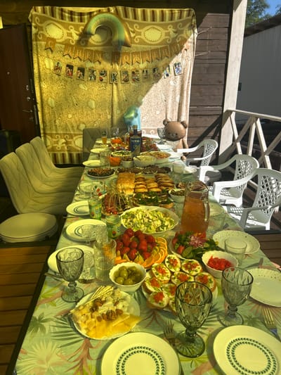 The image shows an outdoor dining table set up on a wooden porch, likely in Lyamino, Russia, during a celebration. A patterned tablecloth covers the table, which is laden with a variety of food and drinks. Visible items include plates, glasses, a pitcher of orange liquid, several bowls of food (some containing olives, strawberries, and what appears to be salad and appetizers), a platter of cheeses and nuts, and several small sandwiches with toppings. In the background, a draped fabric with a shell pattern serves as a backdrop, decorated with a banner that reads "С ДНЕМ РОЖДЕНИЯ" (Happy Birthday) and a string of photographs. A large balloon in the shape of a rainbow and a small inflatable teddy bear are also present. Behind the table and chairs, wooden railings and the side of a building are visible, suggesting a residential or recreational setting. The lighting indicates it is daytime, with sunlight casting shadows on the scene. There are no people clearly visible in the image.