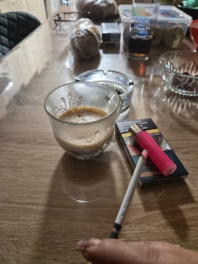 A top-down, close-up shot shows a tabletop with various items on it. A glass of coffee with foam sits centrally, to the left of a partially visible pack of cigarettes with a pink disposable vape on top. A lit cigarette rests on the pack. To the left of the coffee glass are multiple cans of YOVAR Vodka. Further to the left are items in clear plastic packaging, a glass bowl, and a box of "Bapko" baby food. A hand is visible on the right side, holding the cigarette. The table surface has a light brown wood grain pattern. There is no way to determine the location is Stroevo, Bulgaria from the provided image.