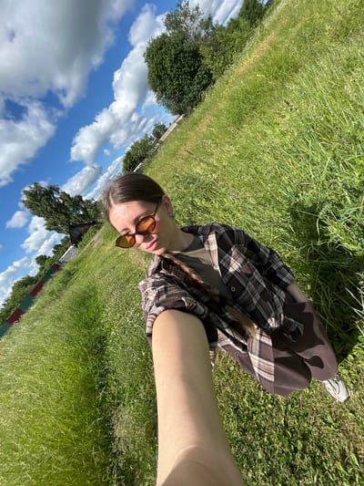 A young woman is taking a selfie outdoors in a grassy field. She is wearing sunglasses, a brown and white plaid shirt over a green t-shirt, and brown pants. Her long brown hair is braided and hangs over her shoulder. The field is covered in tall green grass, with some small white flowers visible. In the background, there are trees and a rustic wooden building. A fence with red and green sections runs along the left side of the field. The sky is blue with fluffy white clouds. The image appears to have been taken on a sunny day. The location is in the city of Malaya Borshchevka, Russia.
