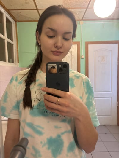 A young female, with light skin and dark hair styled in a braid over her left shoulder, is depicted taking a mirror selfie indoors in Bayronovka, Russia. She wears a white t-shirt with a blue-green tie-dye pattern and a dark graphic design on the front. A simple gold-colored band is on her left ring finger, and small stud earrings are visible. She holds a dark-colored smartphone with a dual-camera system in her left hand; a sticker depicting a small animal is affixed to the phone case near the camera. The background reveals a room with light green upper walls, a light-colored lower wall or tiling, and a panel ceiling. A white panel door with a decorative carving is visible to the right. A white-framed window with multiple panes is to the left. A spherical white light fixture is mounted on the ceiling. The floor appears to be light-colored tile, and a dark, indistinct object is in the lower-left foreground.