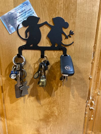 A black metal key holder shaped like two stylized monkeys holding hands is attached to a wooden door. Three sets of keys are hanging from the hooks of the key holder. The leftmost set of keys has several silver keys and a small blue circular object. The middle set has a small golden, textured charm resembling a bag or pouch. The rightmost set includes a black car key fob with a silver emblem that appears to be for an Opel vehicle, along with a couple of silver keys. A business card is partially visible, pinned to the door near the top left of the key holder, with Russian text. The wooden door has a visible wood grain pattern and some minor wear or damage on the right side. The lighting suggests an indoor environment. There is no information to suggest this scene is located in Yokohama, Japan.