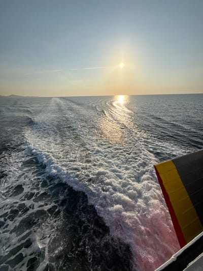 The image displays a maritime scene near Palaiopoli, Greece, observed from the stern of a vessel underway. The sun, a bright orb, is positioned low on the horizon, slightly left of the image center, casting a prominent, shimmering reflection path across the dark blue-gray water. The sky is clear and pale blue, with a faint contrail visible in the upper left. Directly behind the vessel, a substantial white foam wake churns and spreads across the sea surface. A distant, low-lying landmass is discernible on the far left horizon. The lower right quadrant of the image shows a section of the vessel's stern, characterized by dark gray plating with a distinct horizontal yellow stripe and a red lower edge. No individuals or other active vessels are visible. The lighting indicates either sunrise or sunset conditions.