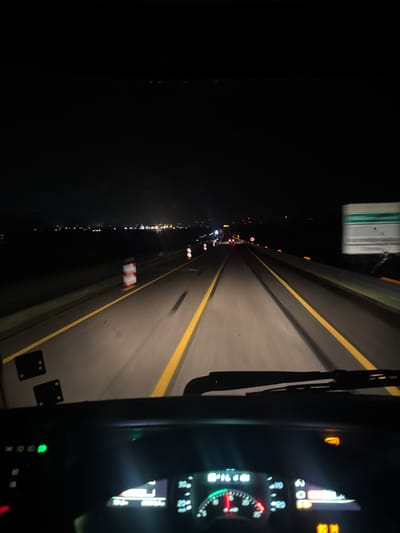 The image captures a nighttime scene from the driver's perspective inside a vehicle on a multi-lane highway near Cerignola, Italy. The roadway is illuminated by the vehicle's headlights, revealing asphalt with prominent double solid yellow lane markings. Distant artificial lights are visible on the horizon, indicating a populated area or industrial complex.
On the left shoulder, a white and red striped traffic bollard is present, suggesting roadwork or a temporary lane boundary. Vehicle lights, including red taillights and white headlights, are discernible in the distance, indicating other vehicles on the roadway. A rectangular green and white road sign is partially visible on the right side of the frame.
The lower section of the image displays the vehicle's dashboard, featuring illuminated instruments. A speedometer with a red needle, a tachometer with a green arc, and several active indicator lights are visible, including a green light on the left and an amber light on the upper right. A black windshield wiper rests at the base of the windshield. The overall environment is dark, with artificial light sources providing the primary illumination.