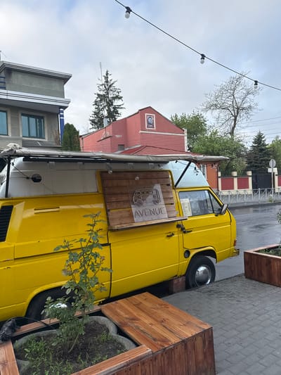A yellow Volkswagen van, converted into a food truck, is parked on the side of a street in Akhieli, Georgia. The van, labeled "AVENUE," has a wooden sign attached to its side and an awning extended. The surrounding environment includes buildings of varying architectural styles, trees, and wooden planters. String lights are visible overhead. The ground appears to be wet, suggesting recent rain. No people are visible.