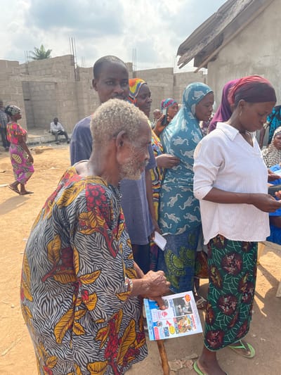 An outdoor scene in Osara Gada, Nigeria, depicts a gathering of approximately ten individuals, predominantly adults, under a hazy sky. The ground is dry, light-colored soil.
In the foreground, an elderly man with a grey beard and short, curly grey hair is seen in profile, facing left. He wears a loose-fitting tunic with a black and white zebra print base overlaid with vibrant red and yellow floral patterns. His left hand holds a folded multi-colored leaflet with text and images, which rests on a dark wooden walking stick.
Directly behind the elderly man, a younger man in a dark blue t-shirt looks directly at the viewer. To the elderly man's right and slightly behind him, a woman wearing a white and blue striped long-sleeved shirt and a green patterned skirt is looking downwards, potentially at an object in her hands. She wears green flip-flops.
Further back in the group, several other individuals are visible. A woman wears a teal hijab with a white cheetah pattern and holds a white piece of paper. Another woman wears a purple head covering. One woman in the mid-ground appears to be holding a dark-colored smartphone to her ear or viewing its screen.
In the left background, a woman in a pink and yellow patterned dress walks away from the camera. Beyond her, an unfinished building constructed of concrete blocks with exposed rebar is visible. A solitary man in a light-colored shirt is seated on a concrete structure near the unfinished building. To the right, another structure with a partially collapsed wooden roof is present. Palm trees are visible in the distant background.