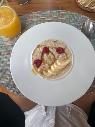 The image shows a white plate with oatmeal, banana slices, and raspberries on top. The plate is on a multi-colored placemat. To the left of the plate is a glass of orange juice. The person who took the photo appears to be wearing white shorts. The location of the photo is a restaurant in Ouled Jelal, Morocco, at latitude 31.663 and longitude -7.872.