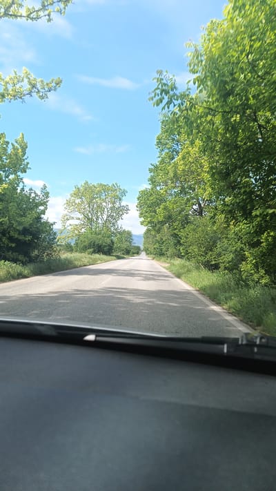 View from inside a vehicle driving on a straight asphalt road in Boliartsi, Bulgaria. The road is flanked by green trees and vegetation on both sides. Distant mountains are visible on the horizon. The sky is blue with scattered clouds. The dashboard of the car is visible in the foreground with a windshield wiper. No people are visible.