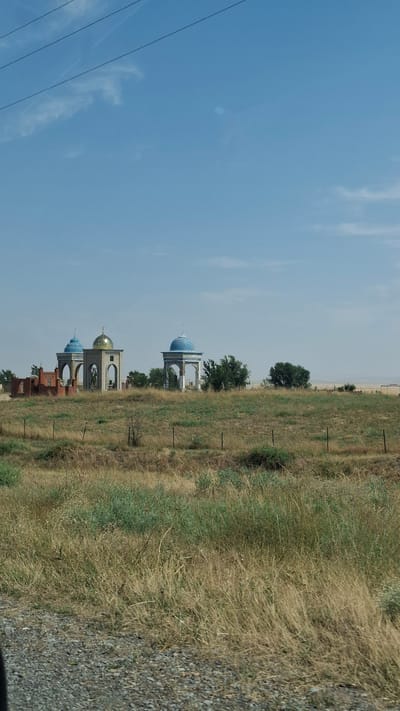 The image shows a row of domed structures located on a grassy hill under a blue sky with scattered clouds. The structures appear to be mausoleums or memorials, with distinct blue and gold domes. In the foreground, there is dry grass and a gravel road. There are no people visible in the image. The location is identified as Tespe, Kazakhstan.