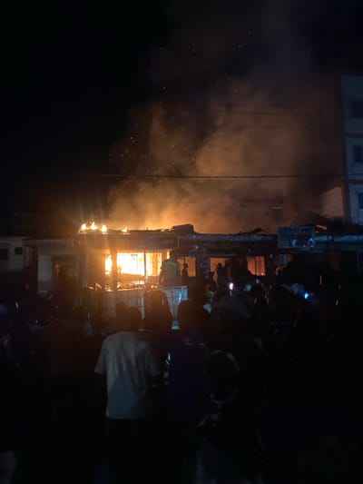 The image depicts a building engulfed in flames, located in the city of Victoria, Cameroon, at the coordinates 4.009 latitude, 9.214 longitude. The structure appears to be a commercial building with a visible storefront. Smoke billows from the roof and windows, indicating a fire in progress. A crowd of people, primarily silhouetted against the blaze, are gathered in front of the building, witnessing the incident. Several individuals are visible in the foreground, some holding flashlights, while others observe the scene. The fire illuminates the night sky with an orange glow, and the environment is smoky and chaotic.
