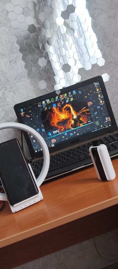 A laptop is positioned on a wooden desk, with its screen displaying a dynamic wallpaper featuring a figure engulfed in flames. The laptop's keyboard is visible, and on the desk to the right of the laptop sits a black and white cylindrical electronic device, possibly a portable charger or speaker. To the left of the laptop, a smartphone is propped up on a white stand, which is part of a circular LED light ring. The background features a textured wall with a pattern of reflective hexagonal tiles and a marble-like texture. The overall scene suggests a workspace or personal setup, potentially for content creation or entertainment. The lighting indicates it is daytime, but no specific weather conditions are discernible. The image provides no clues about the time of day, weather, or location beyond the Bamboura, Abkhazia context. There is text visible on the laptop screen, including the word "easy" and a progress bar with "90%". Other text elements are present in icons and file names on the desktop, but are not clearly legible.
