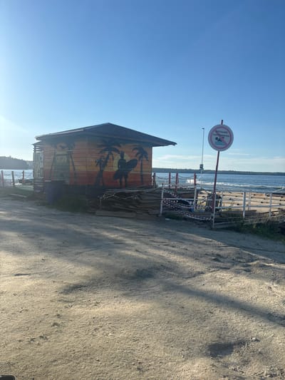 The image depicts a beachside scene on a sunny day. In the foreground, a sandy path leads towards a small, wooden beach hut decorated with palm trees and a surfer silhouette painted in warm sunset colors. Adjacent to the hut, a wooden platform or pier structure is partially visible, cordoned off with red and white caution tape, suggesting it is either under construction or closed. Piles of wooden planks are stacked near the platform.
A prominent circular sign with a red border is mounted on a pole, displaying a pictogram of a person swimming within a red circle with a diagonal line through it, indicating that swimming is prohibited. The text below the pictogram reads "КУПАНИЕ ЗАПРЕЩЕНО!" (SWIMMING PROHIBITED!). Beyond the platform, a body of water, likely a lake or sea, stretches to the horizon, with gentle waves visible. The sky is clear and bright blue. The location appears to be the beach area of Санаторный, Russia, characterized by its waterfront setting and recreational facilities. No people are present in the immediate vicinity.