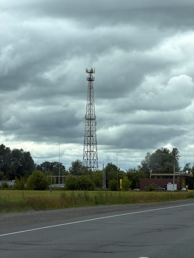 A tall, lattice communication tower stands prominently in the center of the frame, dwarfed by a vast, overcast sky filled with heavy, gray clouds. The tower is a dark, metallic structure with a series of platforms and antennas at its peak. To the left of the tower, a thin, vertical antenna or pole rises from the ground. In the middle ground, a band of green trees forms a dense line, with some sparse vegetation and tall grasses in the foreground. A paved road with a white line marking its center runs across the bottom of the image, suggesting it is a roadside view. Behind the trees on the right, a brown building with a yellow canopy structure is visible, possibly a gas station or service station. A dark-colored car is parked near the building. The overall atmosphere is muted and overcast, typical of a cloudy day in the region. The image is a still, static view with no discernible human activity or interaction.