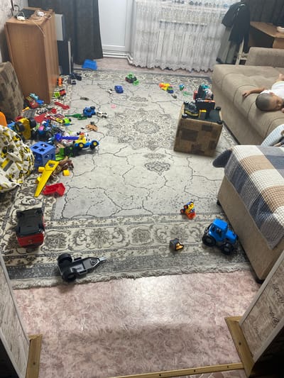 The image depicts a living room in a residential setting, possibly in Бескөл ауылдық округі, Kazakhstan. The room is cluttered with toys, primarily toy vehicles. In the foreground, a red toy truck is overturned on a patterned rug. Further back, a blue toy tractor is positioned near a beige sofa. A child is asleep on the sofa, partially visible. Various other toy cars, trucks, and building blocks are scattered across the rug. The environment appears to be a typical indoor living space with furniture, including a wooden cabinet and a sofa. The lighting suggests it is daytime.