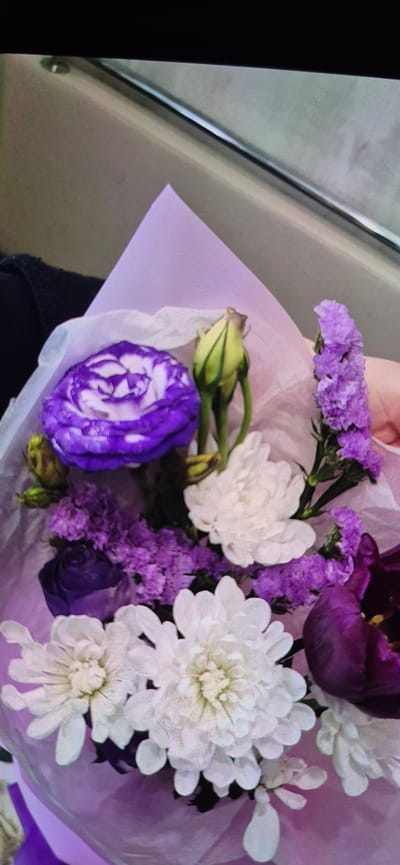 The image shows a close-up of a bouquet of flowers. The bouquet is wrapped in light purple paper and contains a mix of white and purple flowers. Specifically, there are several white chrysanthemum-like flowers, some small purple flowers that appear to be statice, and a larger purple flower with white veining, possibly a lisianthus. There is also a dark purple tulip and a few green buds. The flowers are arranged in a way that suggests they are part of a gift or a floral arrangement. The lighting appears to be indoor, possibly natural light, given the soft shadows and the overall brightness of the scene. The background is blurred, but hints of a metallic surface and a lighter surface are visible, suggesting the bouquet might be resting on a table or another surface in a room. There is no visible text in the image, nor any clear indicators of the time of day or weather conditions. The focus is entirely on the floral arrangement. The setting is likely domestic or a retail space for flowers, given the presentation.