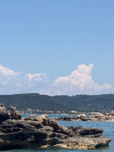 A scenic view of the coast in the Elaties Municipal Unit, Greece, captures a serene seascape under a clear blue sky dotted with fluffy white clouds. In the foreground, weathered, dark grey rocks jut out of the tranquil, clear blue water, with some partially submerged. Behind the rocks, the water continues to a distant shoreline where a strip of sandy beach is visible. A line of buildings, some with red-tiled roofs, are nestled amongst lush green, tree-covered hills that form a backdrop to the scene. The hills extend upwards and are topped with a few scattered trees. The overall impression is one of natural beauty and a peaceful coastal setting.