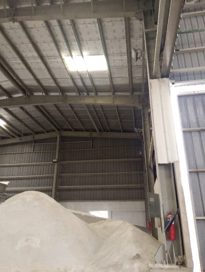 The image shows the interior of a large, industrial-style warehouse or factory in the city of Pangasia, Bangladesh, located at the coordinates latitude 22.694 and longitude 90.601. The building is constructed with a corrugated metal roof and walls.  The roof is supported by a series of metal beams and trusses. A large rectangular skylight in the ceiling provides natural illumination. There is a pile of white powder or sand occupying a large part of the floor space.  A metal pole is visible in the lower right corner of the image.  A red fire extinguisher is mounted to the pole.  A rectangular metal electrical box is also mounted to the pole.  A small window is visible in the lower right corner of the image.  There are no people in the image.  The lighting is bright and even.  There are no apparent signs of damage or distress.