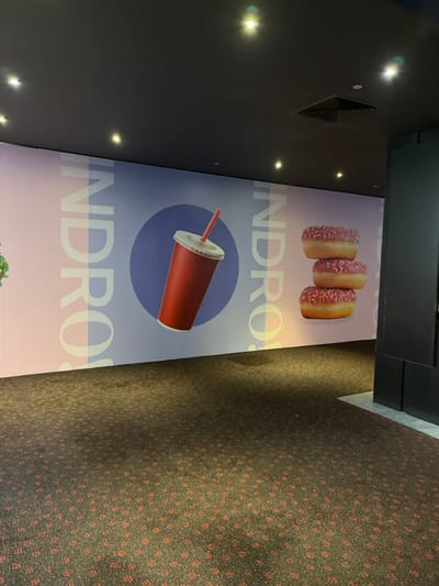 The image shows a hallway with a black ceiling and a large mural on the wall. The mural features a red cup with a straw, a stack of three donuts, and the word "INDRO" repeated twice. The floor is covered in a brown carpet with a red pattern. The hallway is empty and there are no people present. The location is Indooroopilly, Australia, with coordinates -27.501 latitude and 152.972 longitude.  The hallway appears to be a public space, possibly a cinema or a shopping mall. The mural suggests that the area is associated with food and beverage.